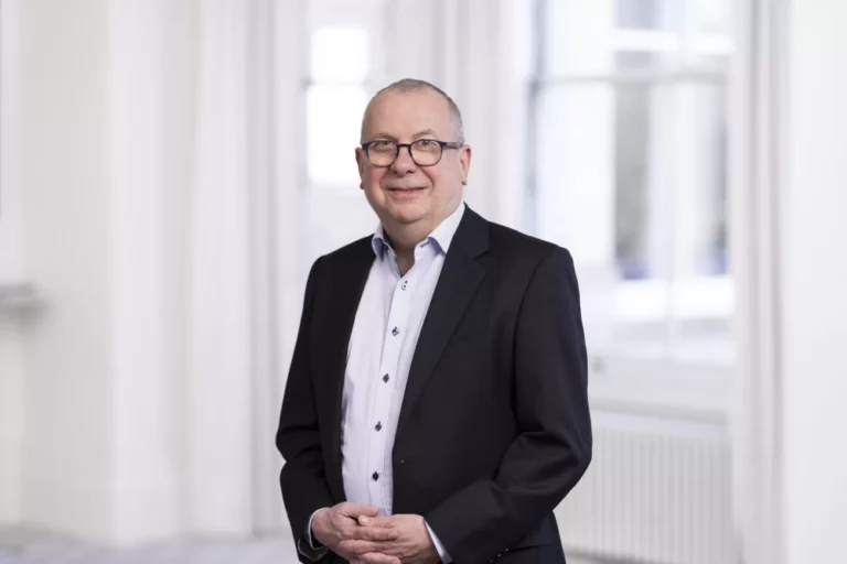 A smartly-dressed man in a suit standing in a brightly-lit white room posing for a photo.