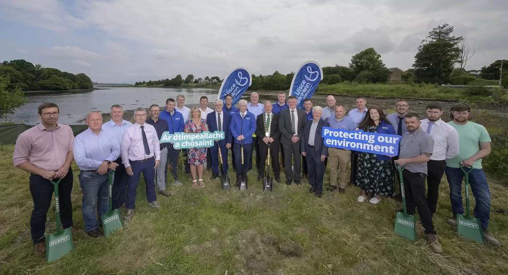 A group of people in business dress stood near a body of water holding signs and green shovels with the Murphy logo.