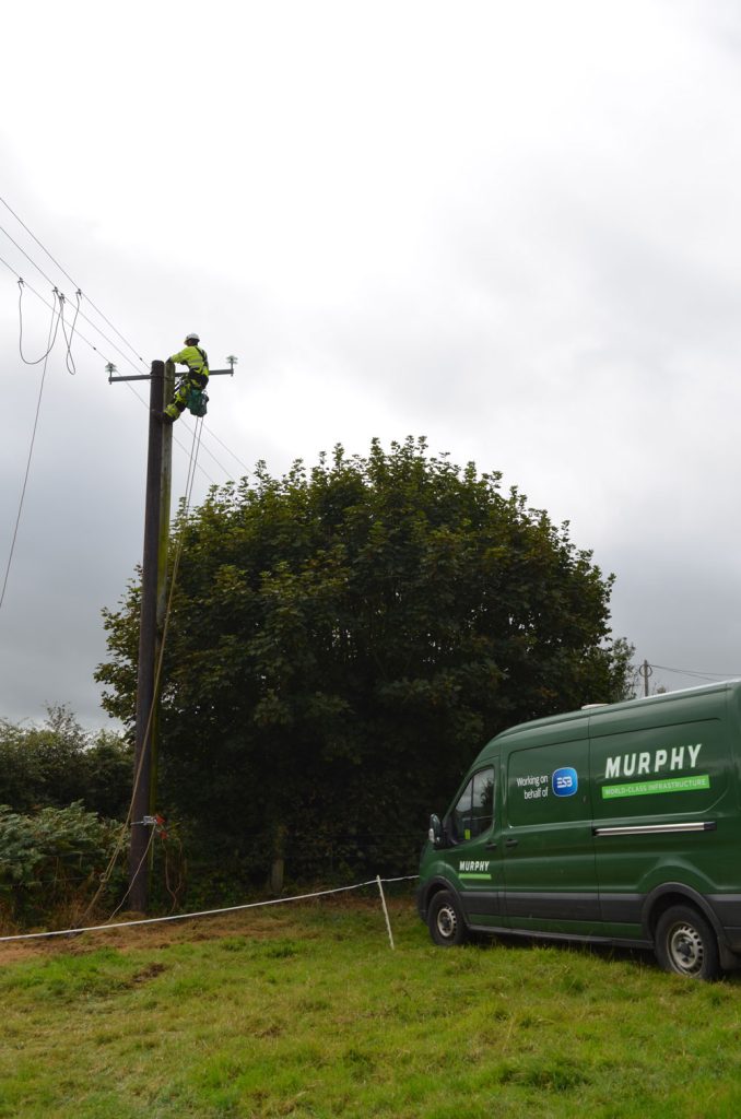 A person in high-visibility clothing working on overhead lines in a field with a Murphy van in the background.