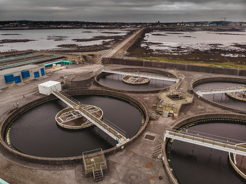 An aerial view of Mutton Island Wastewater Treatment Works connected to a long road surrounded by water with a city scape in the distance