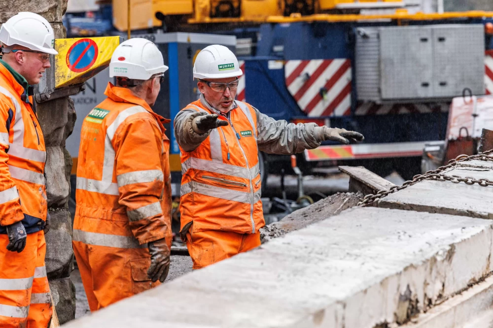 Three people in white helmets and orange high visibility clothing working on a construction site with a construction vehicle in the background.