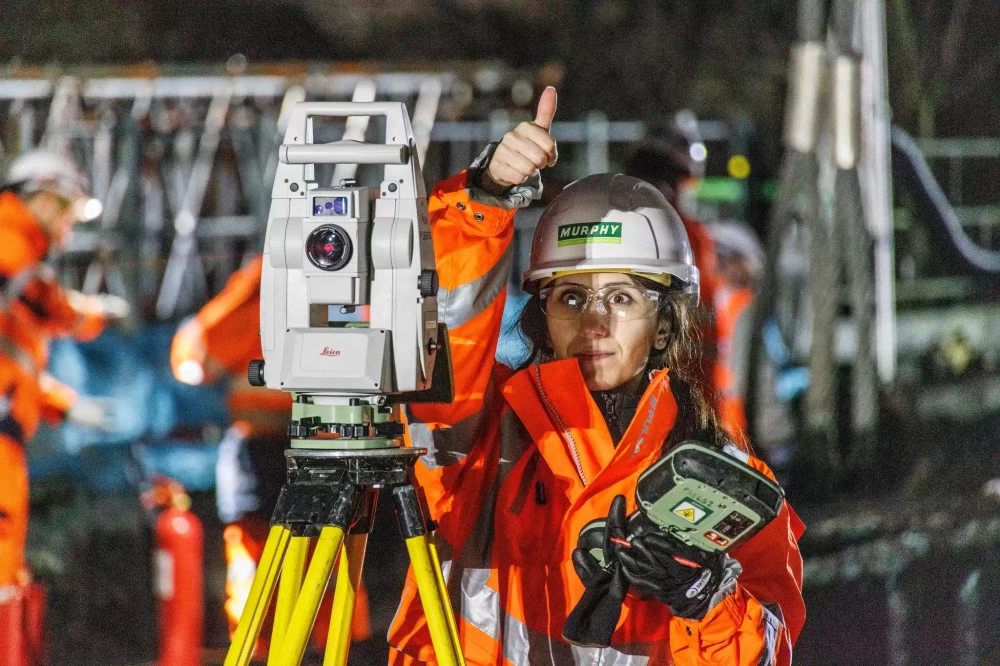 A Murphy employee wearing a uniform and helmet and safety glasses with their thumb up and operating a camera on a tripod.
