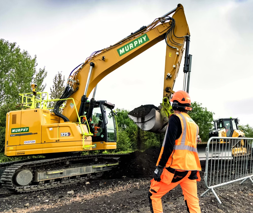 A worker wearing an orange uniform assisting with the operation of a construction vehicle.