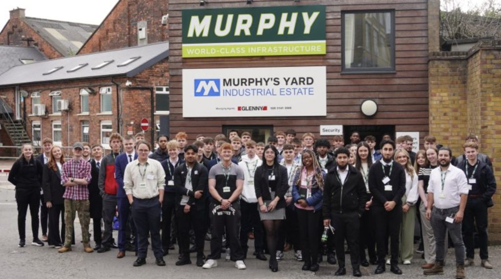 A group of Murphy apprentices stood in front of a wooden building with the Murphy logo and a sign that reads 'Murphy's Yard Industrial Estate'