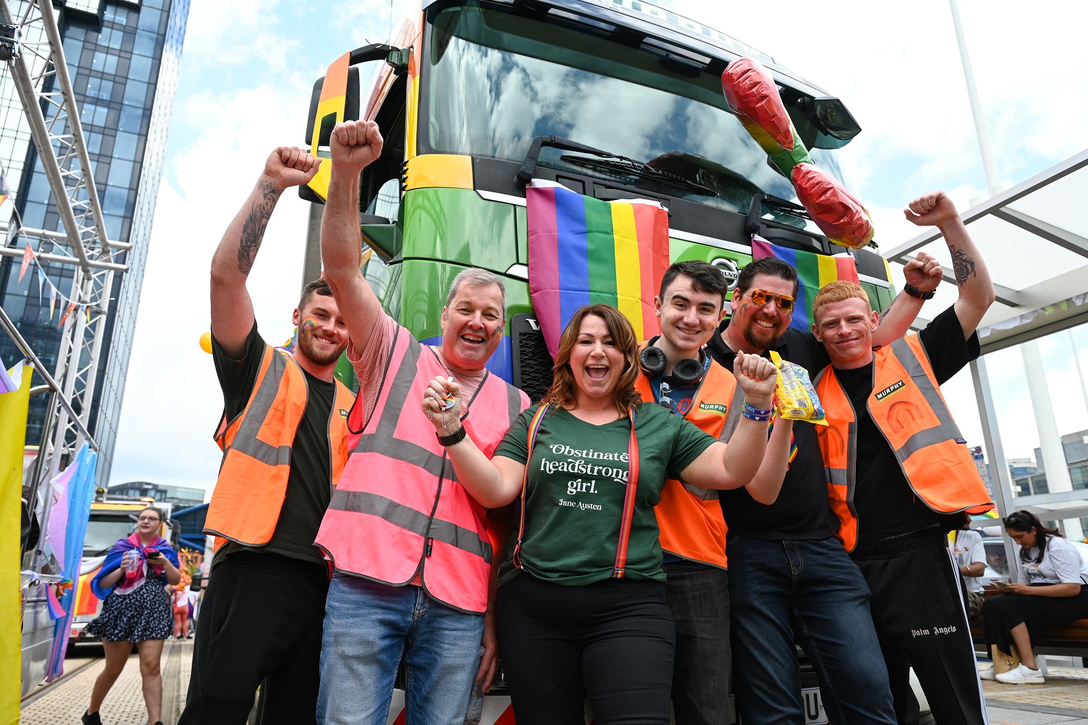 A group of people celebrating wearing high-visibility clothing standing in front of a large lorry with LGBTQ+ flags hanging from the front.