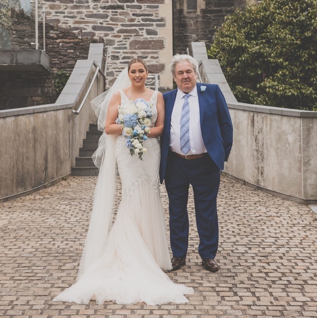 A woman in a wedding dress holding flowers and a man in a suit in front of a historical building.