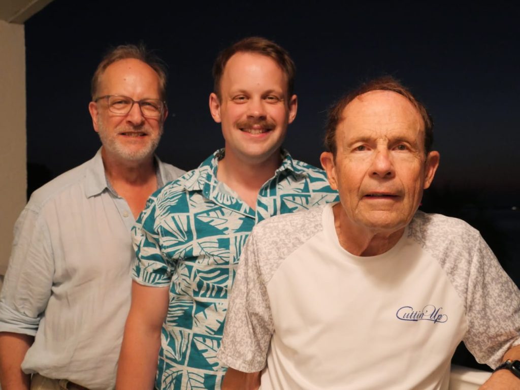 Three men stood in a line wearing bright clothing and smiling.