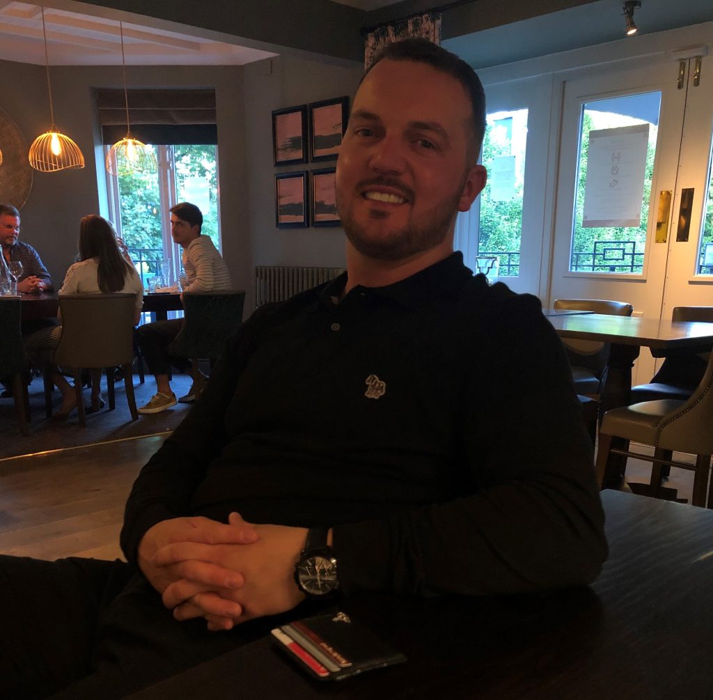 A man sat in a chair smiling with tables and people in the background.