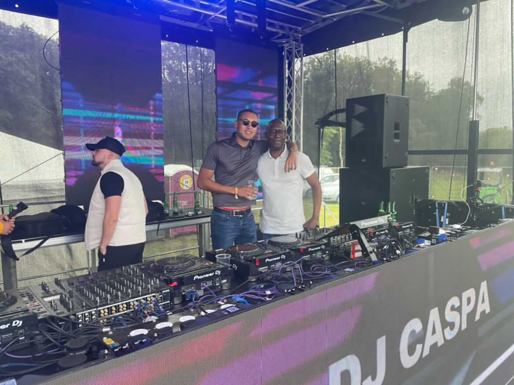 Two men with an arm around each other on a stage stood behind DJ equipment.