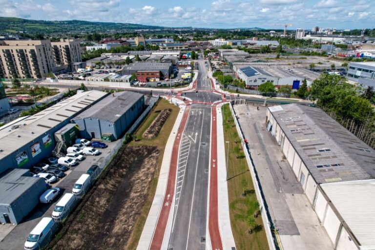 A crossroads from above in the daytime with industrial buildings surrounding it and with hills and a cityscape in the distance.