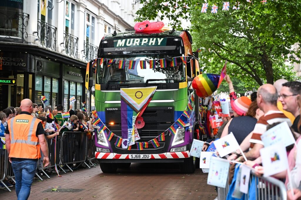 A Murphy branded truck covered in LGBTQ+ flags driving along a street surrounded by people at a Pride event
