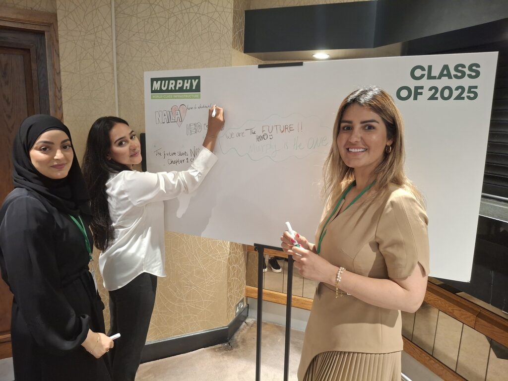 Three women in business dress stood with one writing notes on a whiteboard.