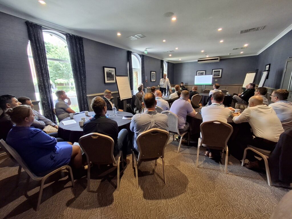 People in business dress listening to a presentation in a brightly lit room.