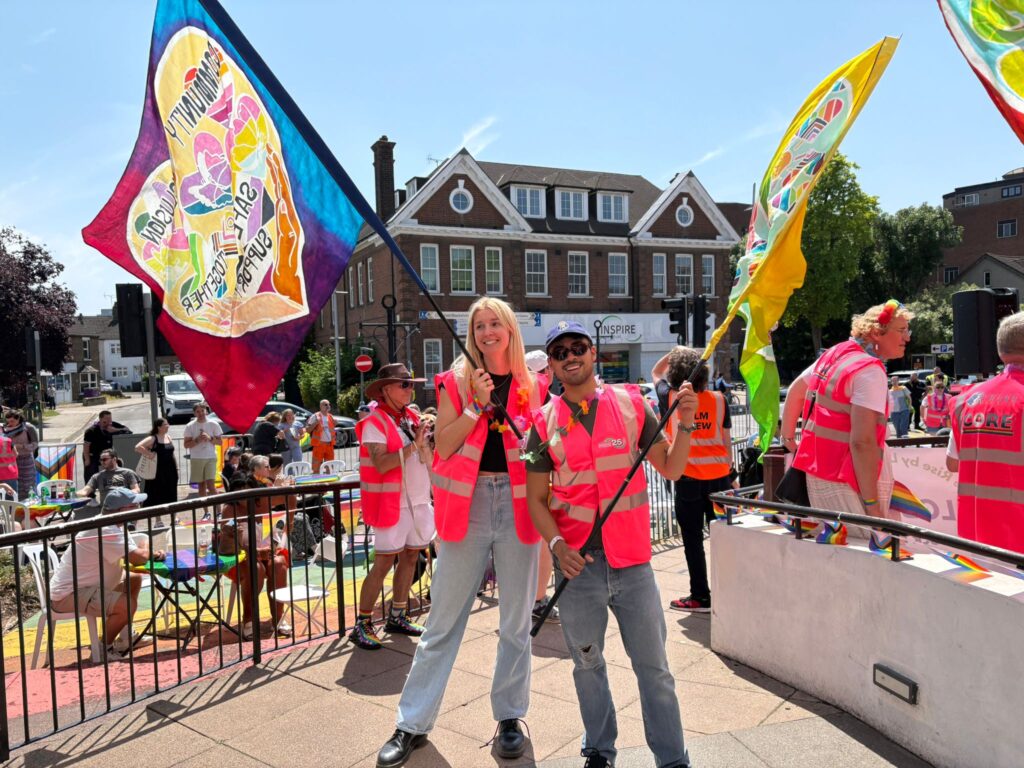 Two people wearing pink high-visibility jackets holding large flags at a Pride event.