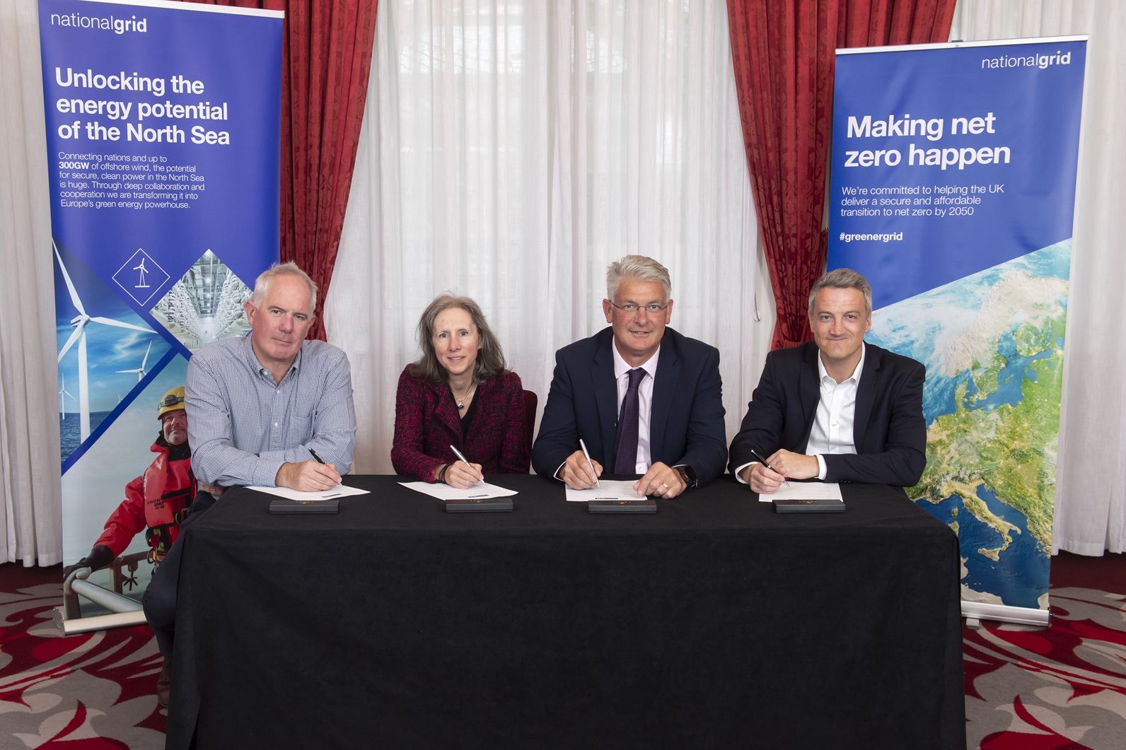 Four people sat a black table signing documents in a room with two national grid roller banners in the background.