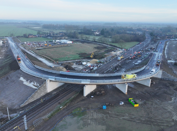 An aerial view of a bridge over a railway track surrounded by fields and trees.