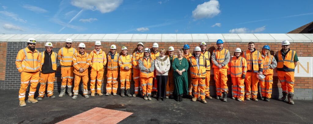 A group of people, some wearing safety uniform and helmets, stood in front of a wall smiling.