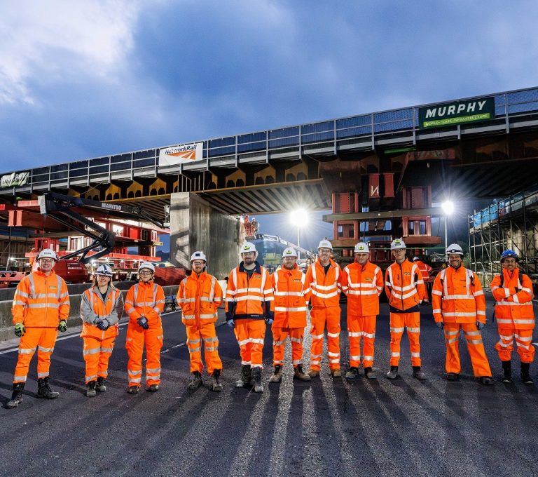 A group of people in high-visibility Murphy uniforms standing in front of a railway bridge under construction.