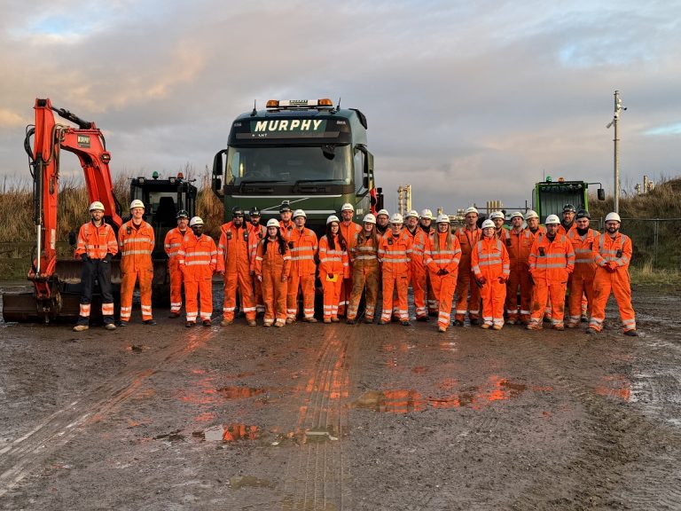 A team of people wearing orange Murphy uniform stood in a line at a construction site in front of a Murphy lorry and a construction vehicle.