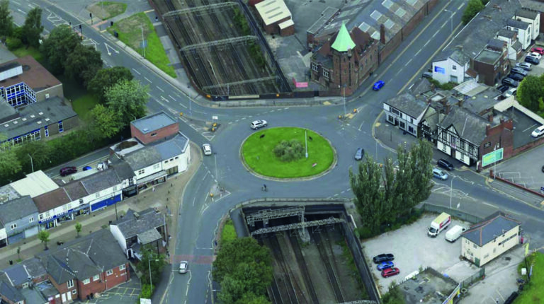 An aerial photo of a urban area in the daytime including a roads, a roundabout, cars and train tracks.