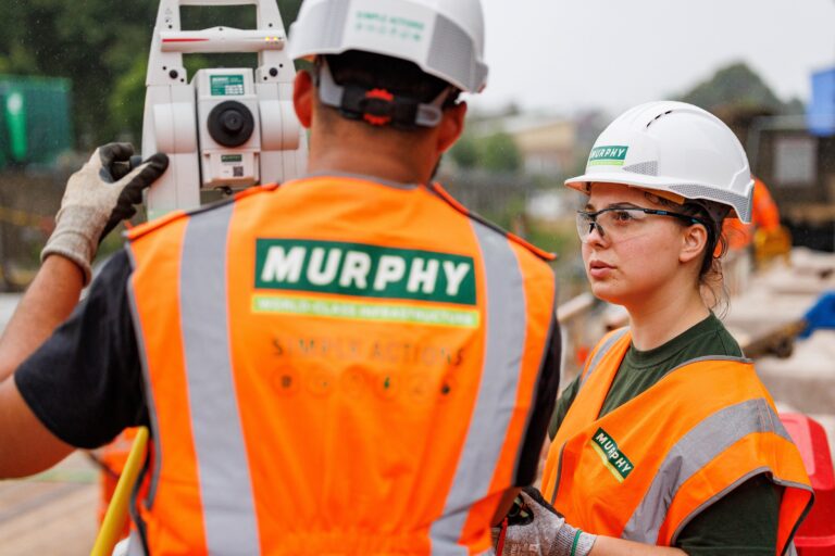 Two people in Murphy high visibility jackets and helmets on a construction site operating a piece of equipment.