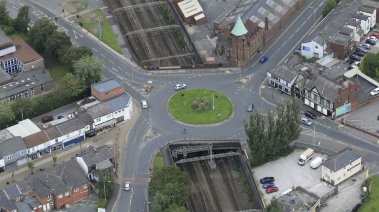 An aerial photo of a urban area in the daytime including a roads, a roundabout, cars and train tracks.